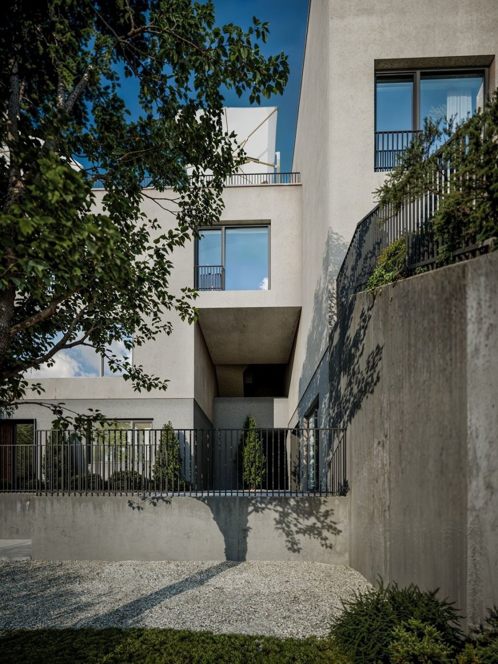Neubau Attika-Maisonette am Heidenweg - Multi-story residential building with white exterior walls, large windows, and balconies. The building is surrounded by trees and has a gravel pathway leading to the entrance.
